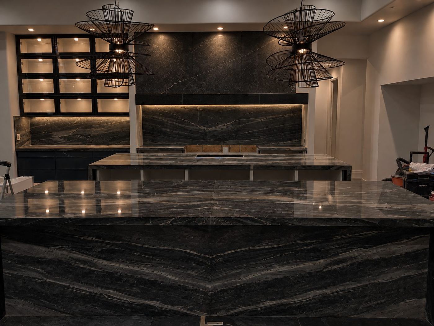 Kitchen with two large islands in dark stone with cream veining, a waterfall edge, and a full-height stone backsplash behind the range